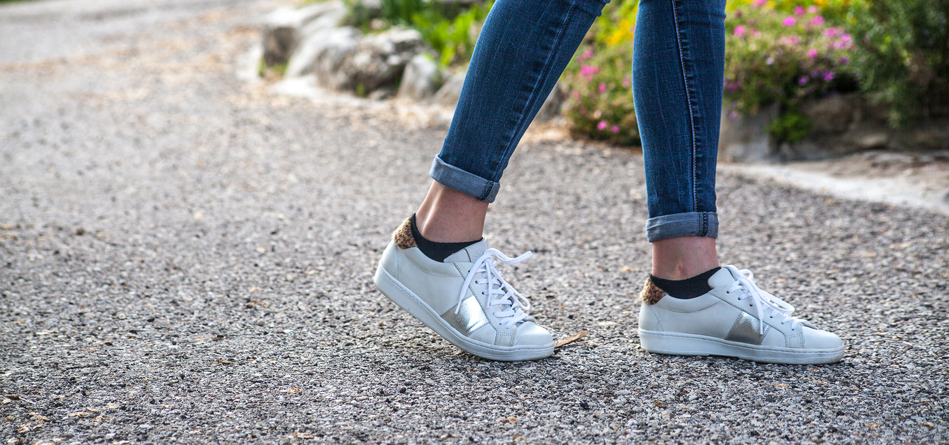Gambe con jeans e sneakers bianche camminano su un sentiero di ghiaia