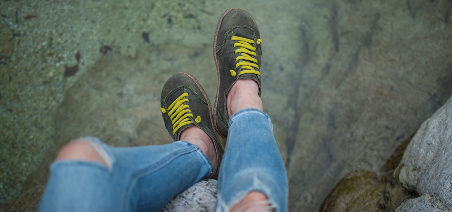 Scarpe verdi con lacci gialli su sfondo acquatico, indossate con jeans strappati.