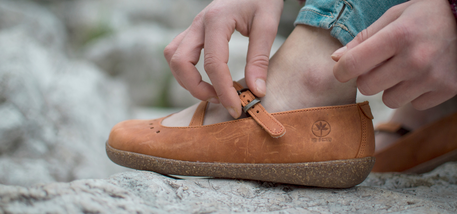 Mano che allaccia cinturino su scarpa cuoio chiaro con logo inciso.