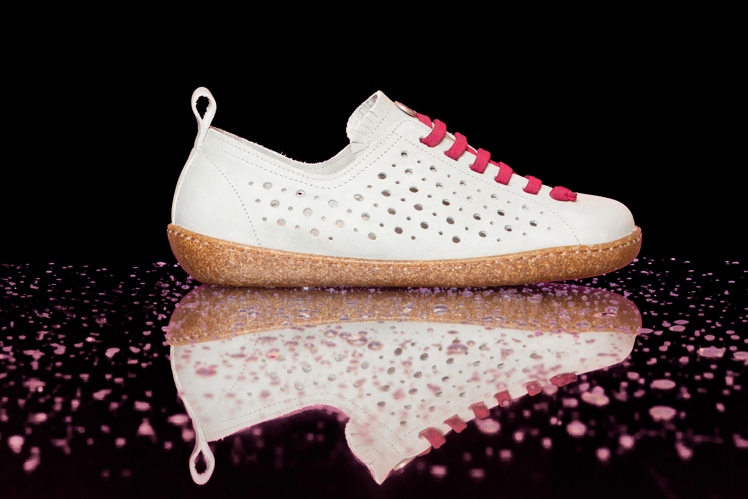Sneaker bianca traforata con suola in sughero e lacci fucsia su sfondo nero lucido.