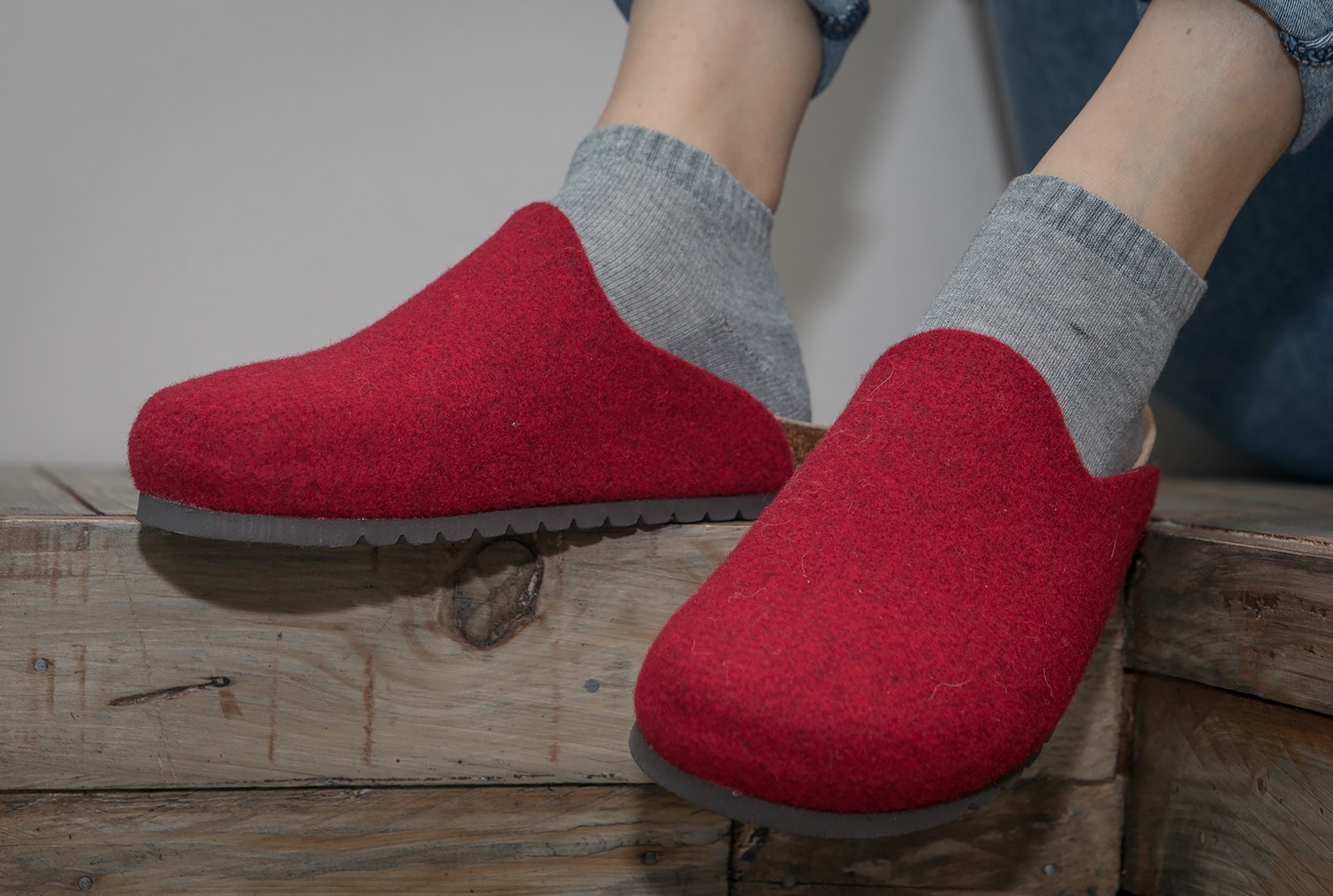 Piedi con sabot rossi in feltro e calzini grigi su asse in legno.
