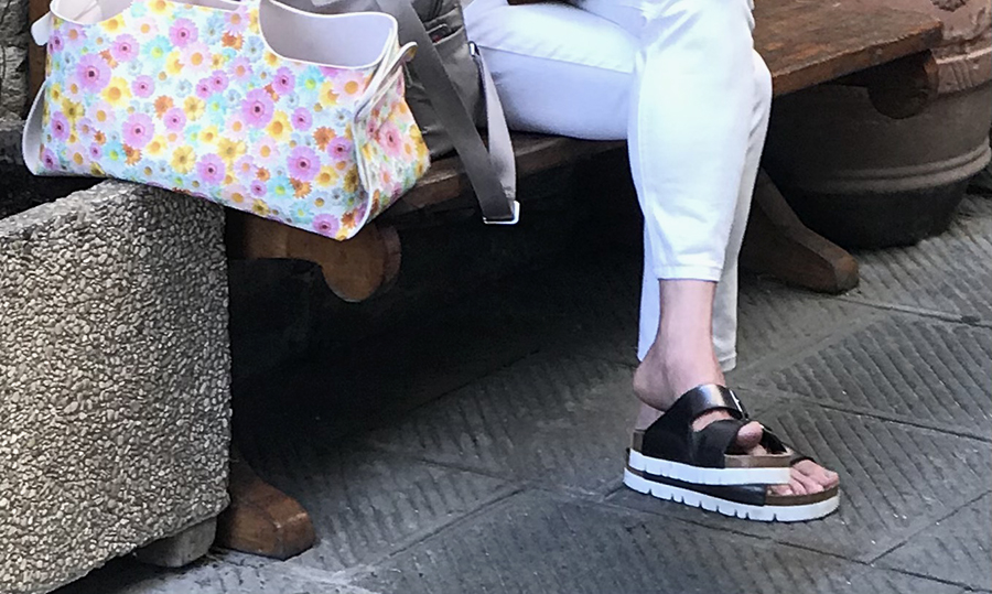 Donna seduta su panchina con sandali platform neri e pantaloni bianchi.