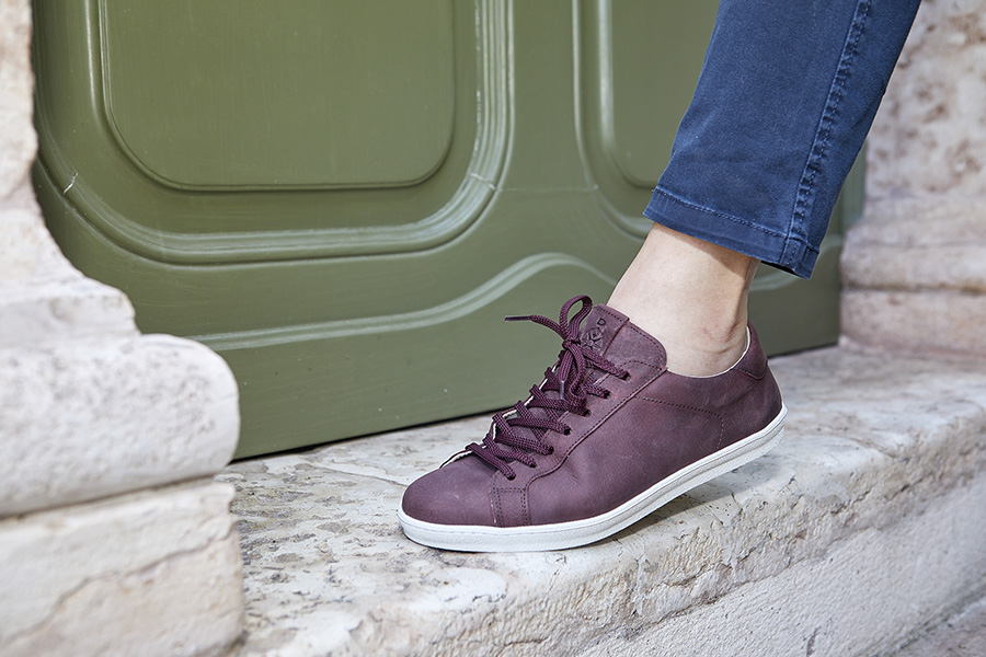 Sneaker bordeaux opaca con suola bianca indossata davanti a una porta verde.