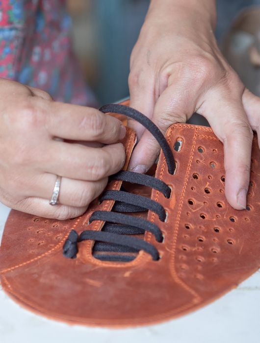 Hands tying black laces on a brown leather upper during the creation of a shoe.