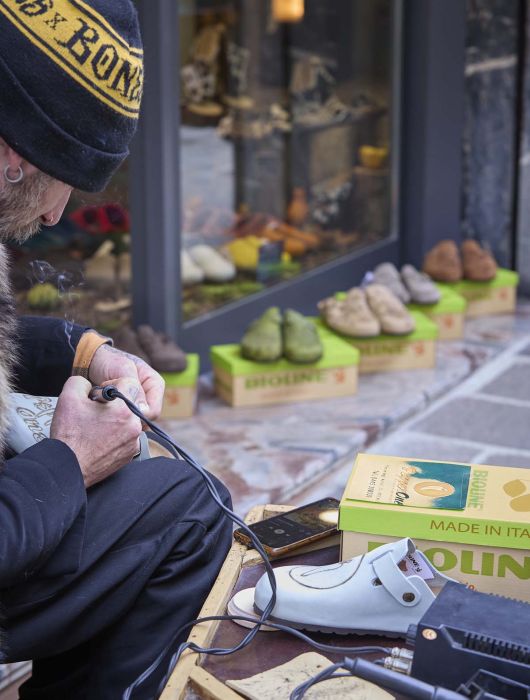 Un uomo lavora su un sabot bianco fuori da un negozio con scarpe colorate esposte in scatole.