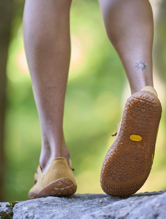 View from below of the legs of a person walking on a rock outdoors, wearing sand-coloured minimalist shoes with rubber soles, in a green natural environment.