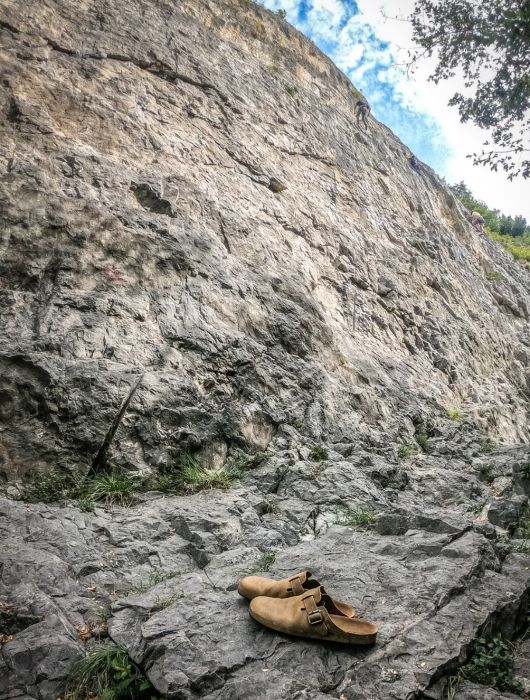 Sabot Bioline cuoio su rocce ai piedi di una parete, con scalatori visibili in arrampicata.